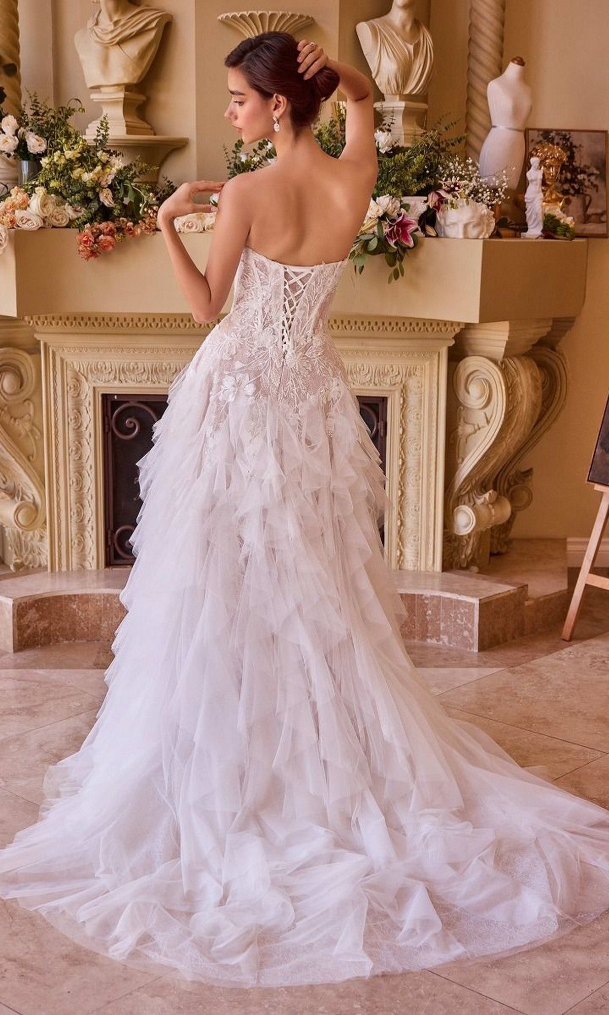 A woman wearing the Andrea and Leo WL016 long wedding dress, featuring a strapless lace-applique corset bodice and ruffle tulle A-line skirt, stands with her back to the camera before a decorated mantelpiece.